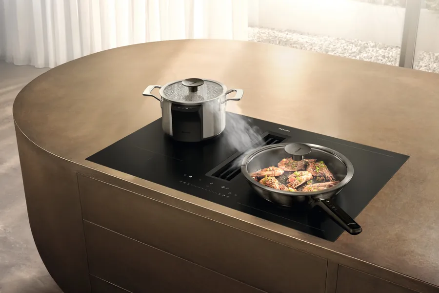 Miele KMDA 8683 FL Induktionskochfeld mit integriertem Dunstabzug in einer bronzefarbenen Kochinsel, Dampf zieht in den mittigen Abzug
